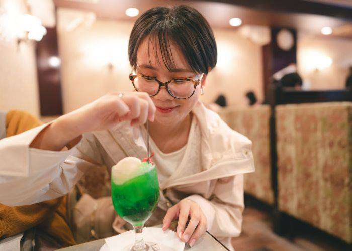 A young lady wearing glasses digging her spoon into a melon soda float.